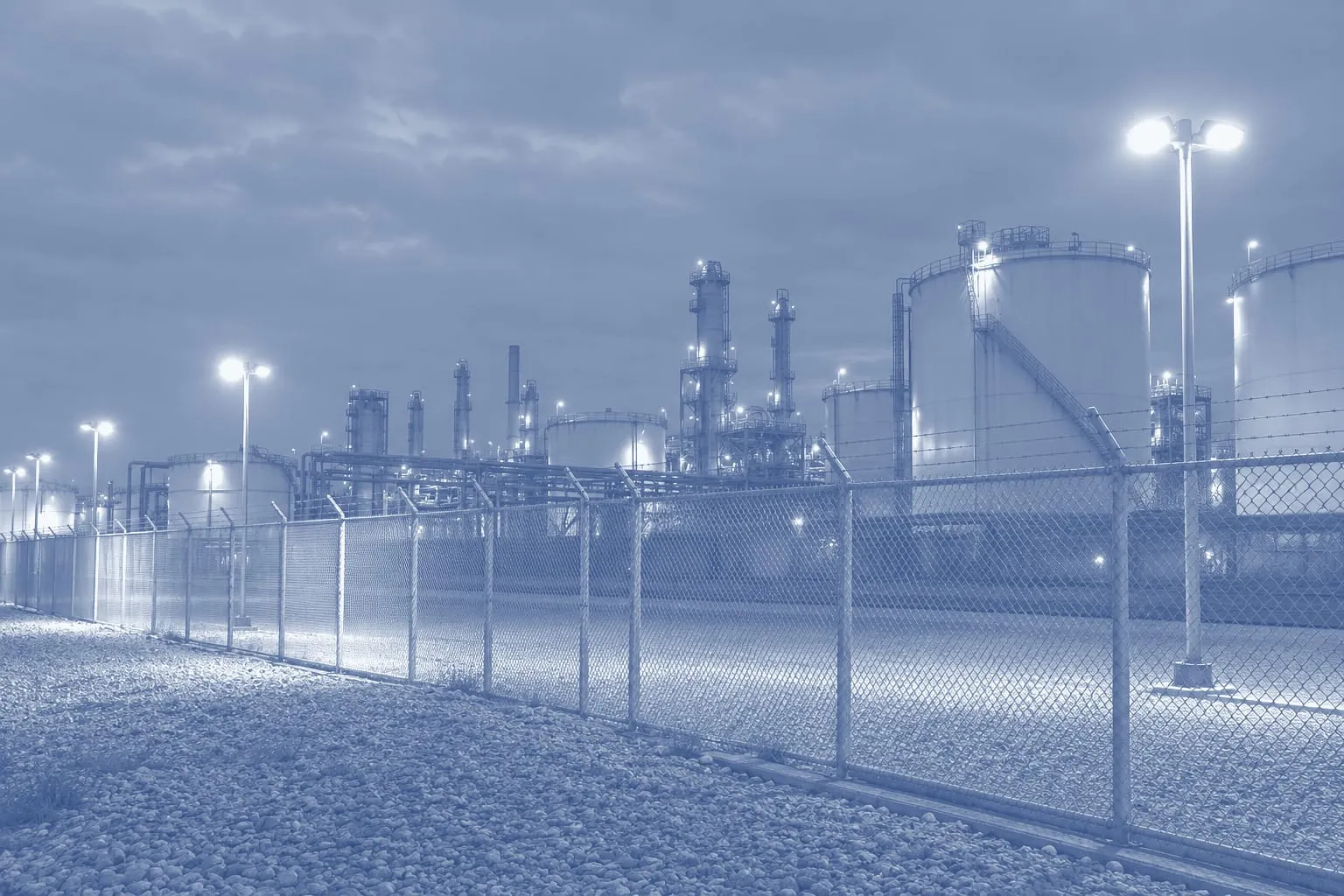 The floodlit perimeter fence of an industrial plant at night, tanks and pipes in the distance, an illustration printed in blue duotone